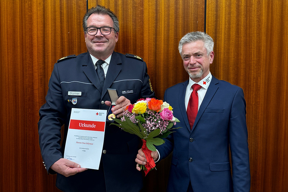 Zwei Männer in Anzügen stehen nebeneinander, der linke hält eine Urkunde und einen Blumenstrauß.