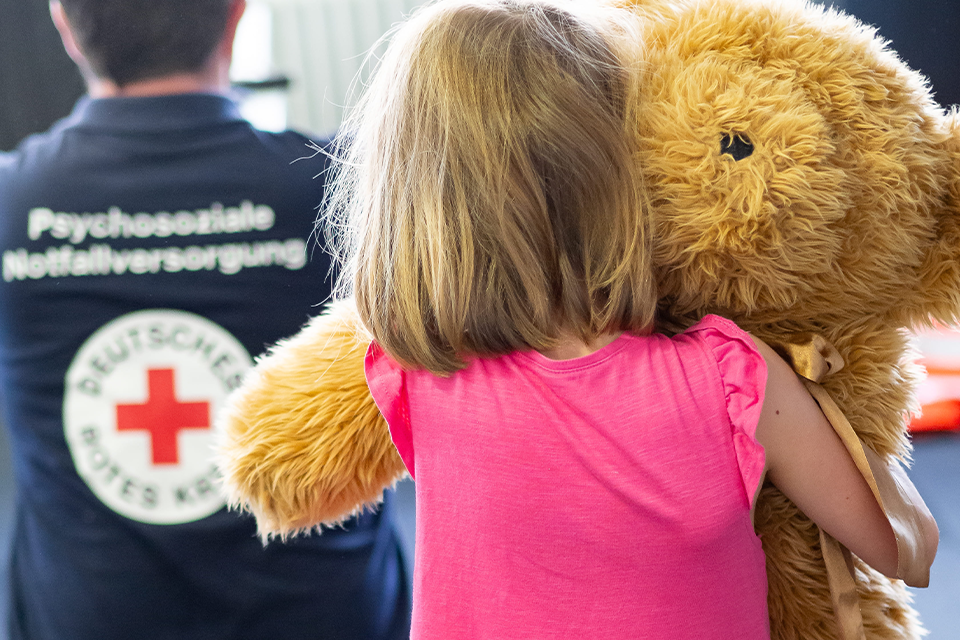 Kinder-Krisen-Einsatzdienst Die Rückansicht eines Kindes mit Teddy, daneben steht ein DRK-Mitarbeiter