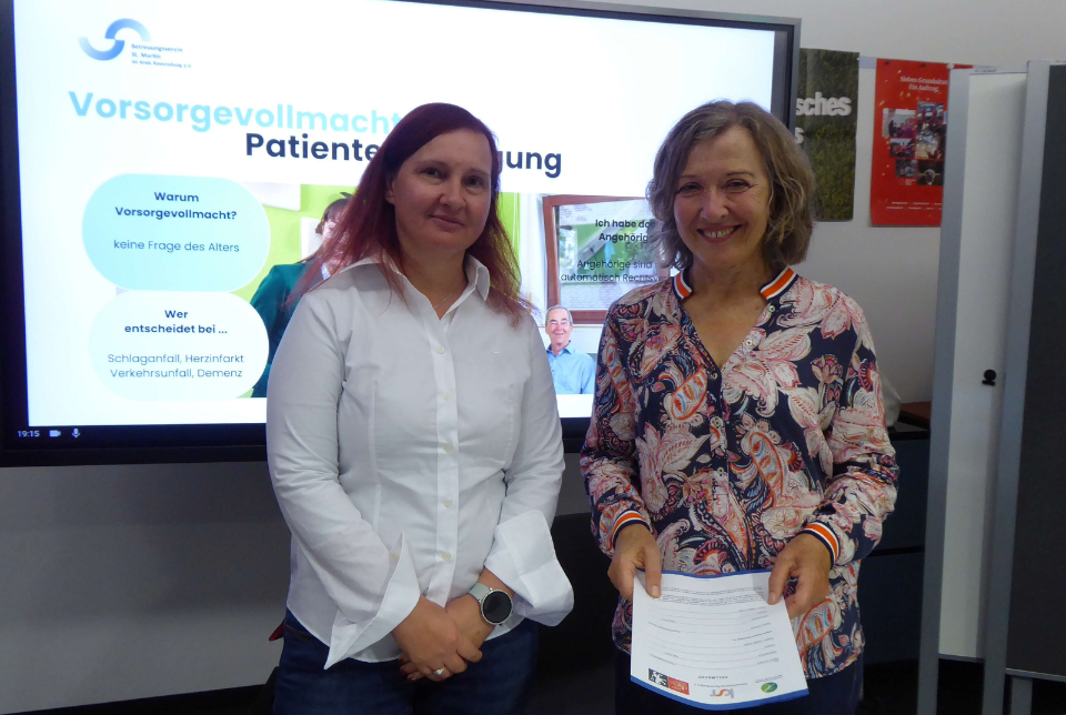 Vorsorge Zwei Frauen stehen bei einer Veranstaltung vor einem Info-Bildschirm